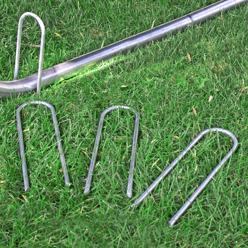 3 wind stakes lay in the grass while 1 wind stake is sunk into the ground over the trampoline leg. 